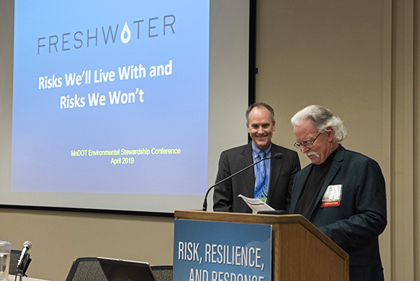 Picture of two men near a podium. One is reading from notes, and the other is looking on, smiling.